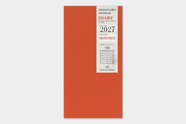 A vertical, orange-red traveler's planner for 2027 with a white paper label on the front showing '2027 Monthly' and 'Traveler's Notebook' text, photographed against a light gray background.