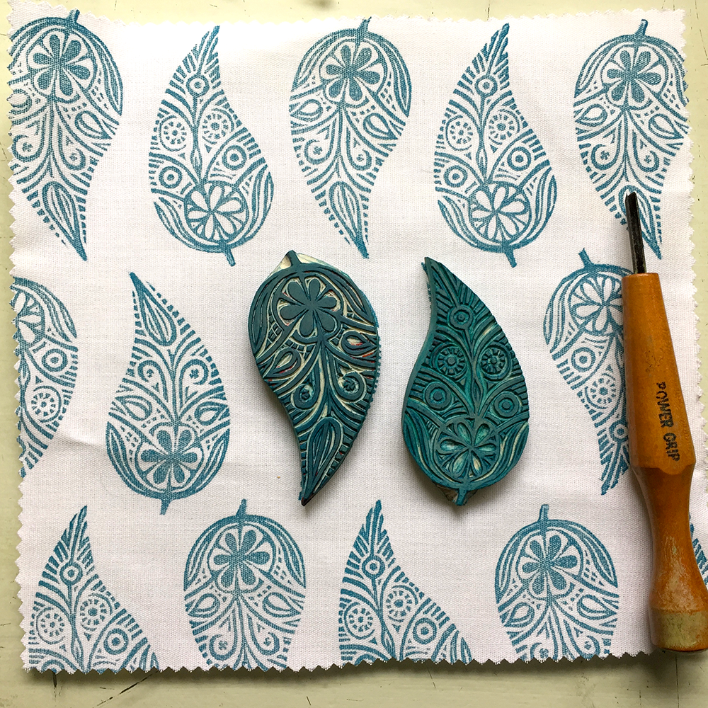 Two carved leaf-shaped carved blocks with teal printed patterns displayed on a white fabric with a wooden carving tool.