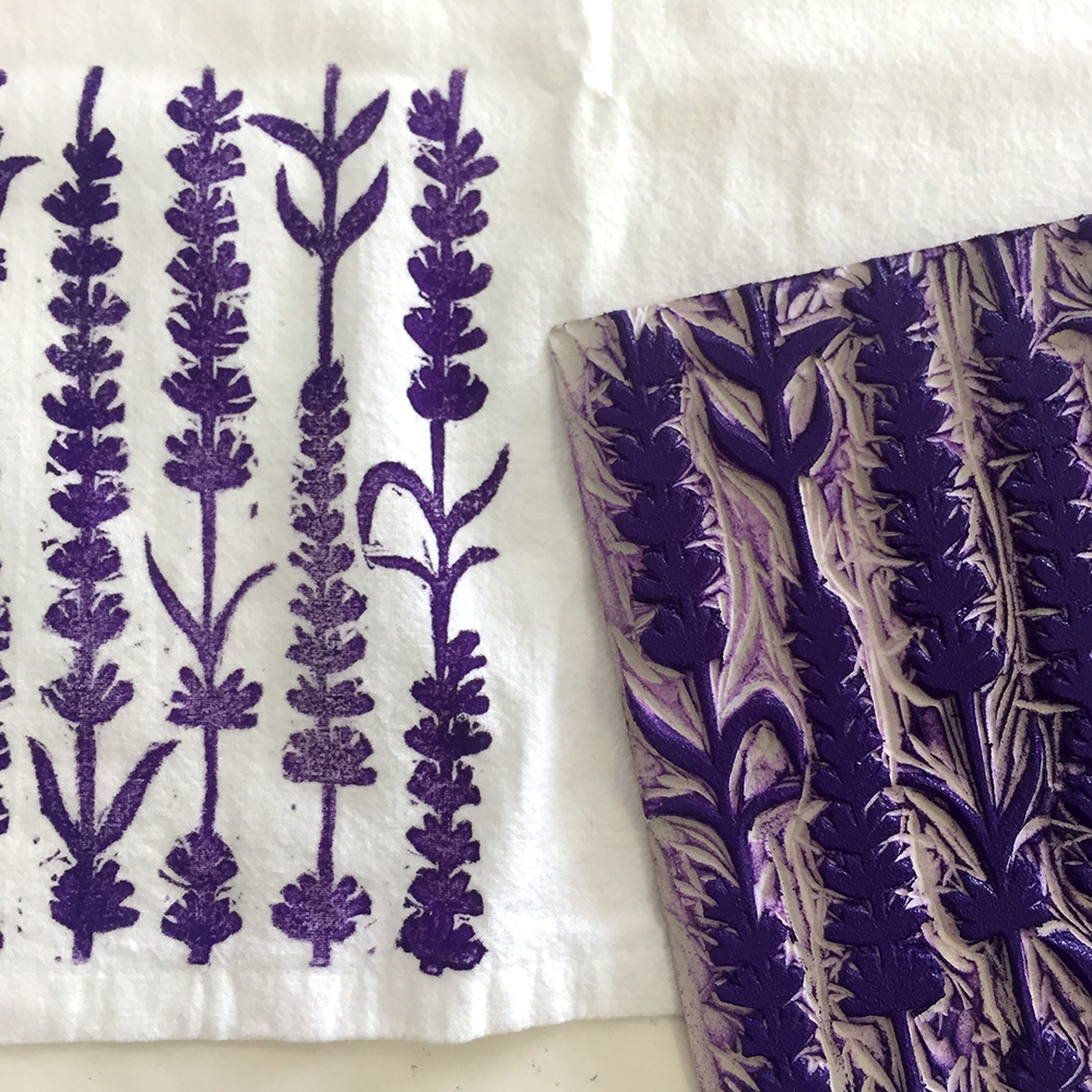 A close-up of a white fabric featuring a purple lavender floral block print. A matching purple and off-white patterned printing block with the same pattern  is placed on top of the printed textile.