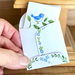 Hand holding a small folded card with a blue bird illustration on a wooden surface