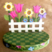 Decorative wooden garden scene with flowers and a white picket fence on a checkered base.