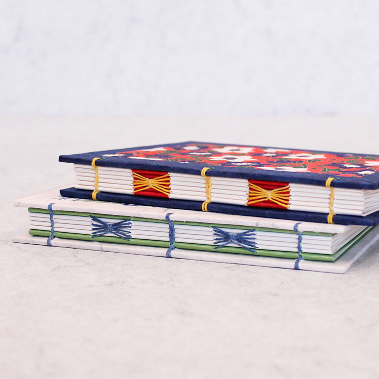 Two decorative handmade paper notebooks stacked on a light surface. The top notebook has a navy blue cover with a red and white floral pattern and yellow cord binding.