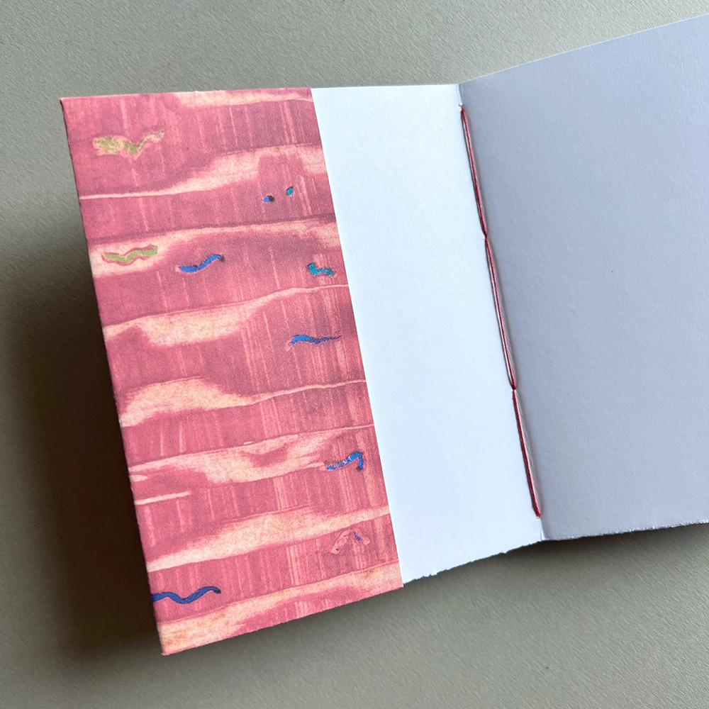 An open journal lying on a neutral gray surface, showing a redpatterned cover with abstract horizontal brushstrokes and small blue and gold accents, alongside blank white pages.