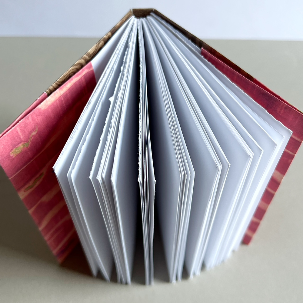 An open handmade accordion-style journal with white pages fanned out, bound with a pink patterned cover and a brown patterned spine, photographed on a neutral beige surface.