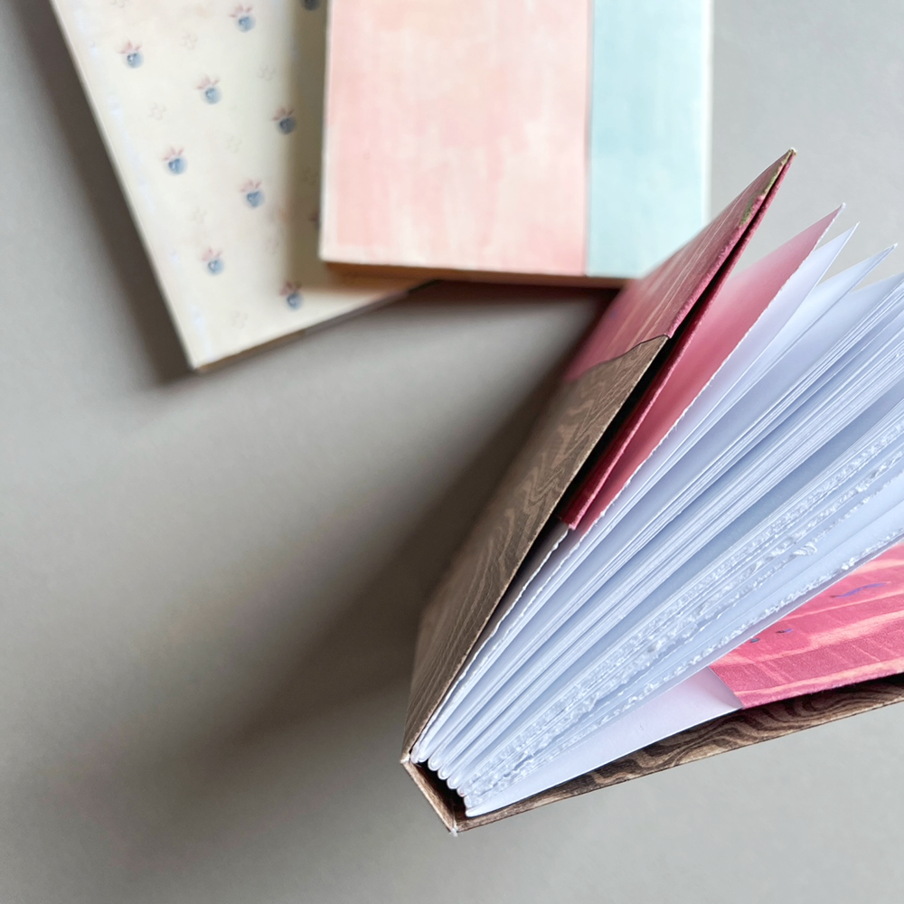 A close-up of an open journal showing a brown wood-grain cover, white pages, and pink and blue patterned interior sheets, photographed on a neutral gray surface.