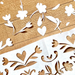 White floral paper cuts laid out on a textured brown wood background, showing cutouts of flowers, leaves, and hearts