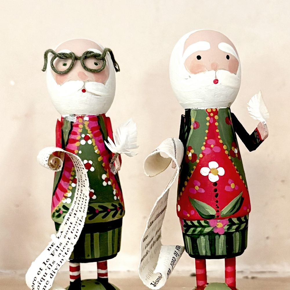 Two hand-painted wooden figurines of Santa Claus with glasses and a long paper list, standing against a plain beige background.