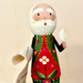 A handcrafted wooden Santa Claus doll with a white paper beard and hat, holding a rolled paper list, dressed in a red and green patterned coat with a white flower on the front, set against a plain beige background.