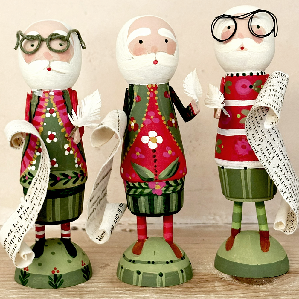 Three hand-painted wooden Christmas Santa Claus figurines standing on round bases, each holding a scroll, set against a plain beige background.
