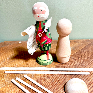 A wooden santa figure with a painted face, floral dress, and rolled paper list stands beside an unfinished wooden peg doll on a wooden tabletop. Several wooden sticks and a round wooden piece are arranged in front against a pale green wall.