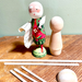 A wooden santa figure with a painted face, floral dress, and rolled paper list stands beside an unfinished wooden peg doll on a wooden tabletop. Several wooden sticks and a round wooden piece are arranged in front against a pale green wall.