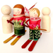 Handcrafted wooden peg dolls dressed in winter clothing, with one doll wearing red and green patterned clothing and holding a ski pole, accompanied by two plain wooden peg dolls on a white background.