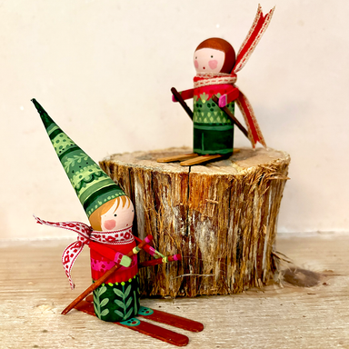 Two handmade wooden gnome skiers displayed on and around a rustic tree stump. One gnome is standing on skis in the foreground wearing a green patterned hat and red outfit, while the other sits on the stump in the background wearing a green sweater and red scarf.