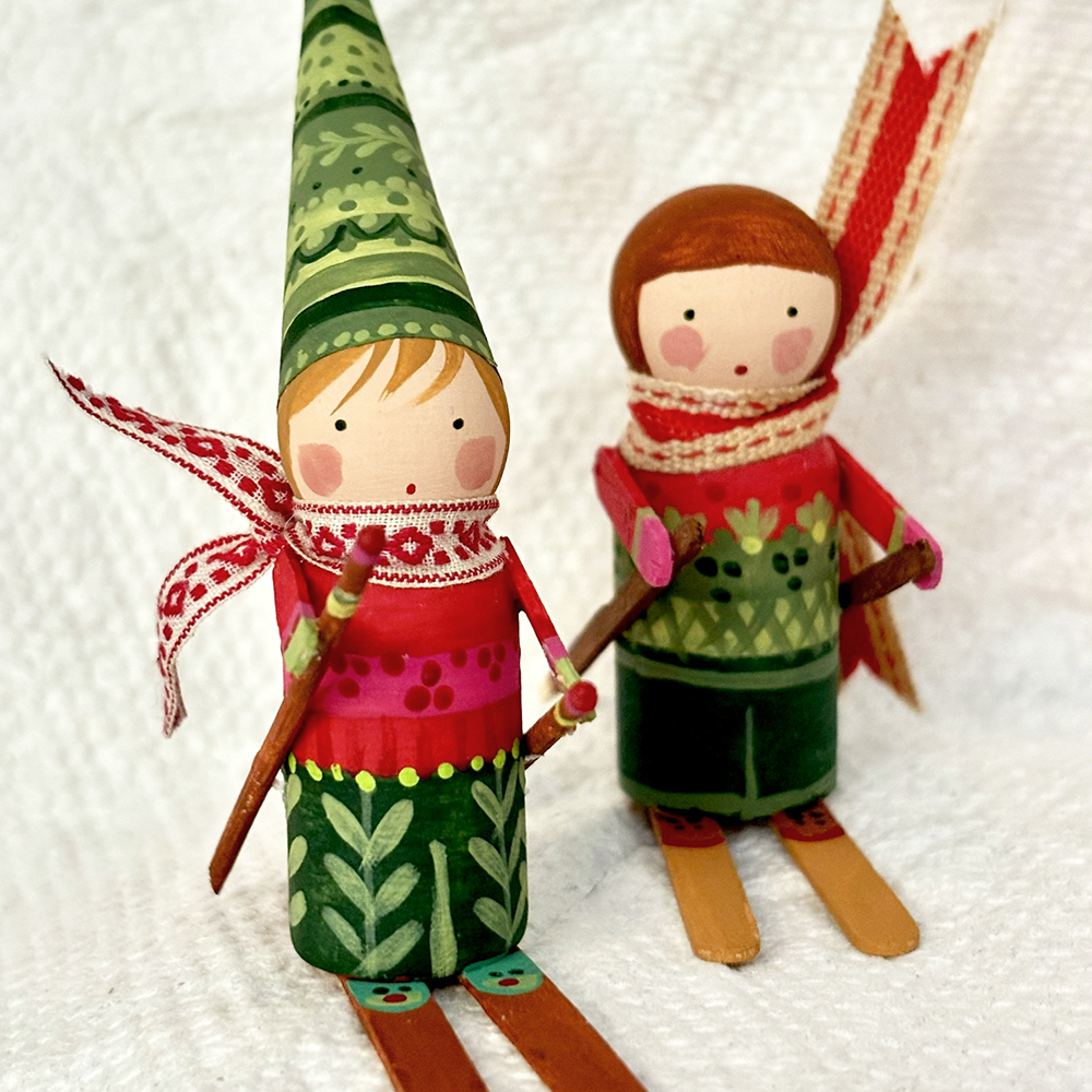 Two handcrafted wooden gnome dolls dressed in colorful winter clothing, standing on wooden skis against a white textured fabric background.