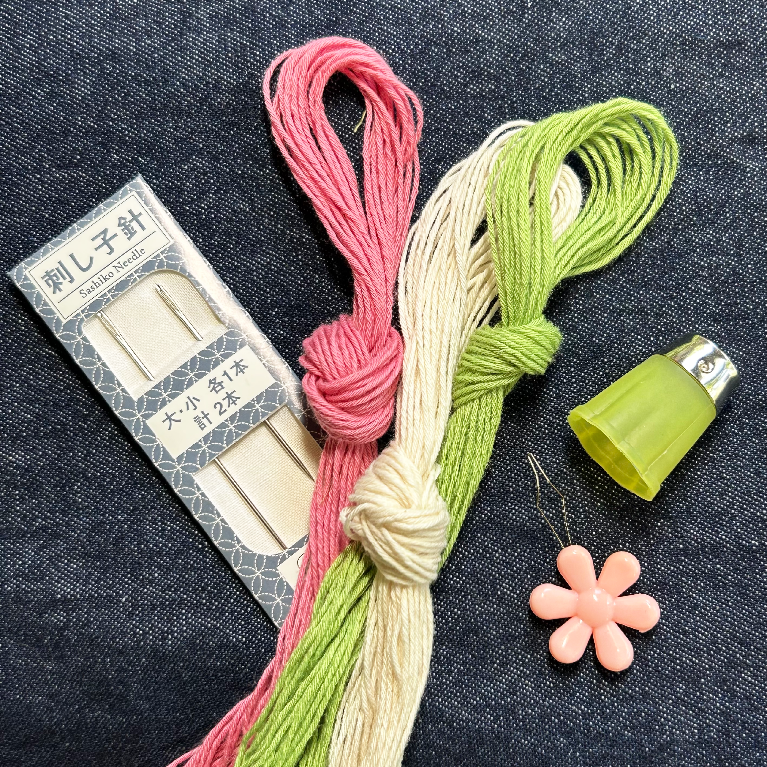 Japanese embroidery thread set with pink, cream, and light green threads, a box of needles, a green thread cap, and a pink flower-shaped stitch holder on a dark denim background.