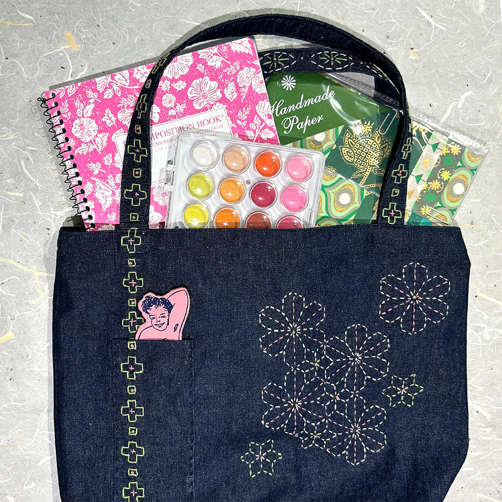 A dark indigo denim tote bag with white embroidered floral patterns and a pink patch,  shown filled with various colorful stationery and craft items on a textured gray background.
