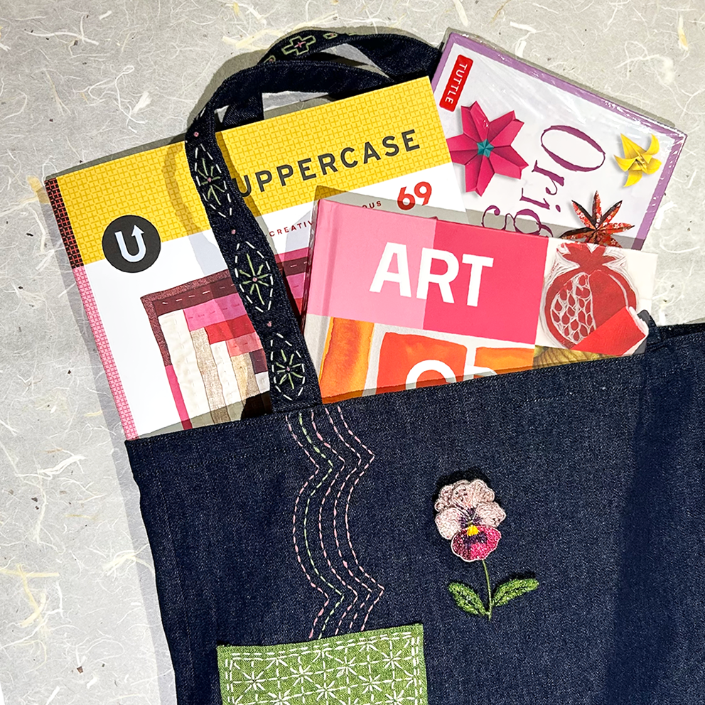 A dark indigo denim tote bag with embroidered floral details and a green patterned pocket, shown filled with colorful books and magazines on a textured light gray surface.