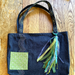 A handmade dark indigo denim tote bag with a green embroidered patch and a multi-colored ribbon tassel, displayed on a wooden surface.