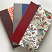 A stack of three hardcover journals fanned out on a light neutral background. The covers feature various patterns and colors,  a floral orange and white pattern, a gray linen-textured cover, a blue polka-dot cover, and  floral paisley pattern with red, blue, and green details.