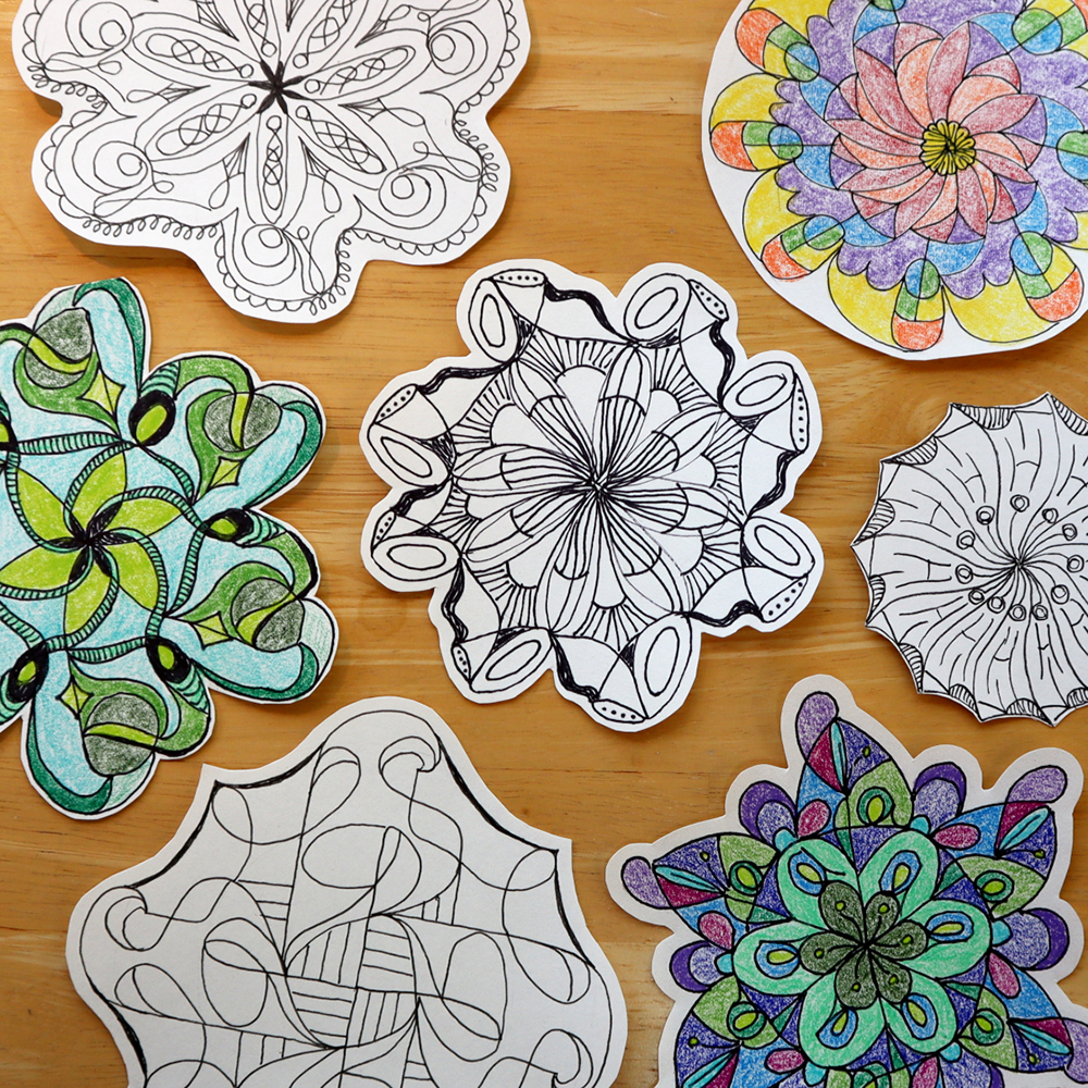 A collection of cut mandala designs displayed on a light wooden background. Some mandalas are black and white line art, while others are colorful and filled with shades of green, blue, purple, pink, and yellow.